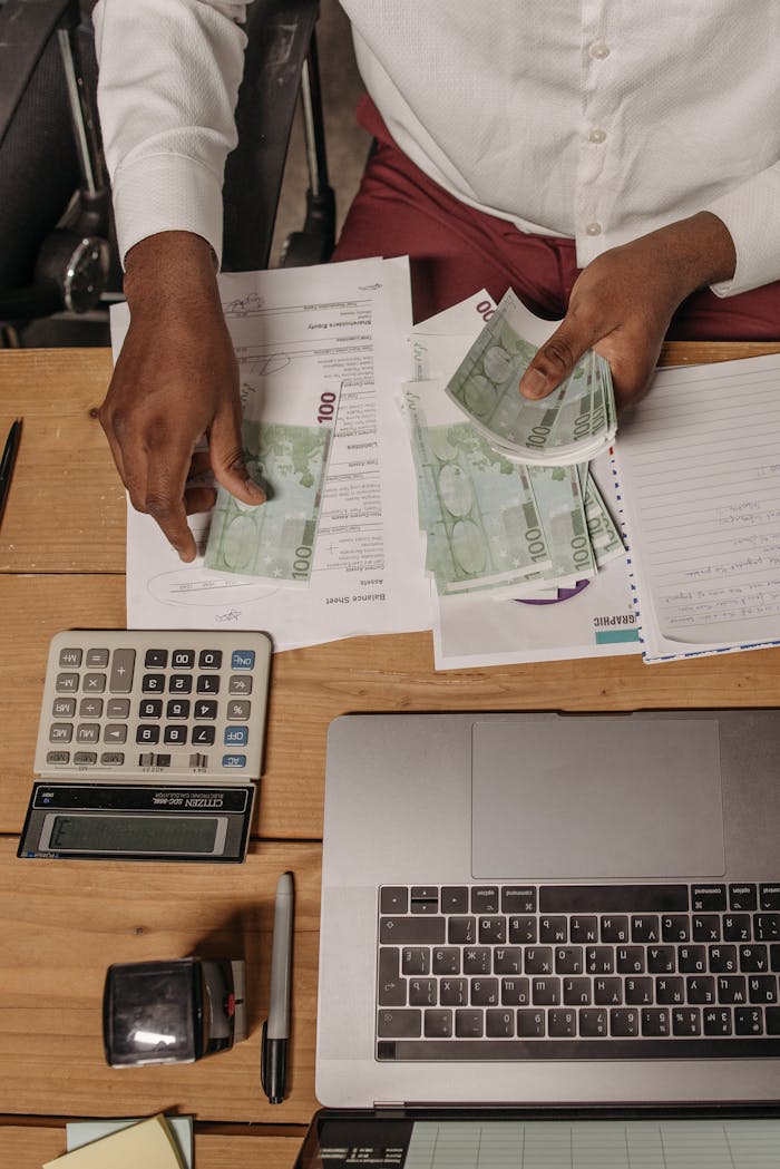 An overhead view of a professional counting cash and using a calculator to manage finances on a wooden desk.